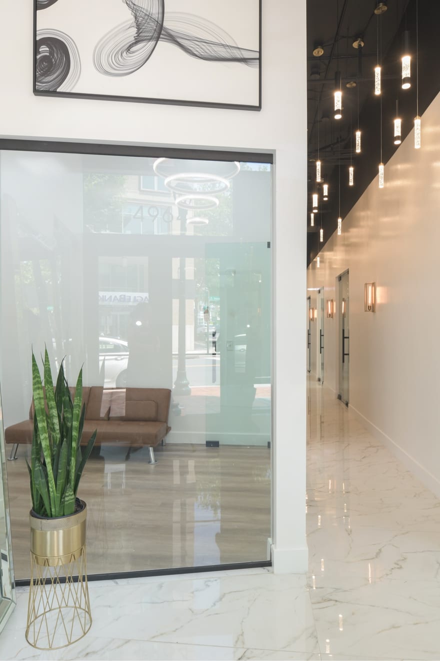The lobby at 4964 Fairmont Avenue: a glass entrance wall, a brass planter, abstract artwork, and the lit pendant corridor running away to the right over a calacatta floor.