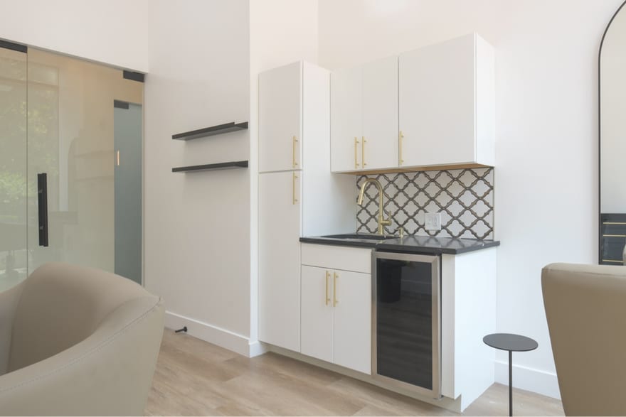 Inside a suite: white cabinetry with brass handles, a black stone counter with a shampoo sink and brass mixer tap, a patterned backsplash and a glass-fronted beverage fridge.