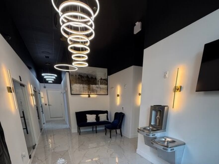 The lobby at 7917-B Westpark Drive: a spiral chandelier of lit rings dropping from a black gloss ceiling over a polished white marble floor, with brass sconces, navy velvet seating and the suite doors running away down the hall.