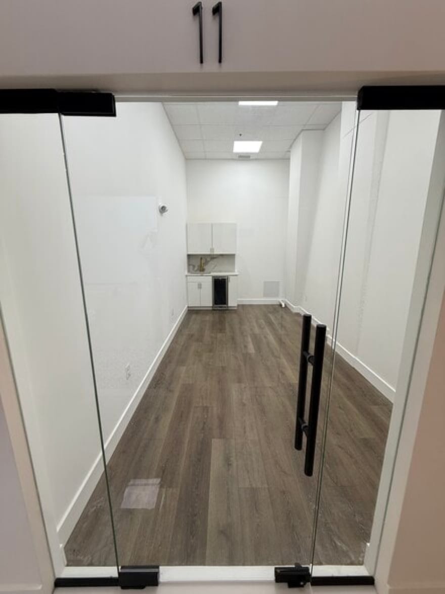 A suite at Westpark Drive seen through its open glass doors, photographed empty before move-in: a room with wood-look flooring, white walls and a white kitchenette with a sink and an under-counter fridge at the far end.