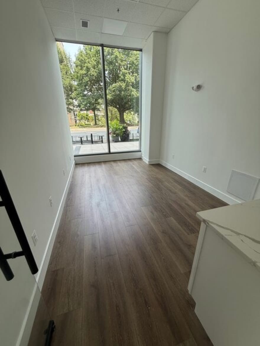 An empty corner suite with a full-height window looking out to trees and the sidewalk seating outside, wood-look flooring and a stone-topped counter in the foreground.